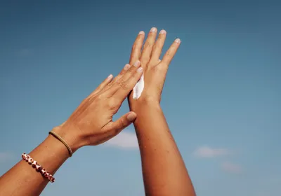 A person applies sunscreen to their hands against a blue sky.