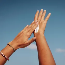 A person applies sunscreen to their hands against a blue sky.