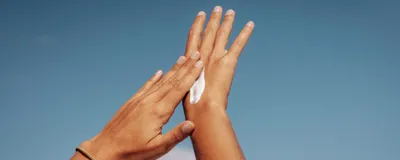 A person applies sunscreen to their hands against a blue sky.