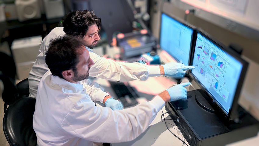 Gabriele Casirati, seated closer to the camera, and Andrea Cosentino point to a computer screen. They are wearing lab coats and gloves. Gabriele Casirati, seated closer to the camera, and Andrea Cosentino point to a computer screen. They are wearing lab coats and gloves.