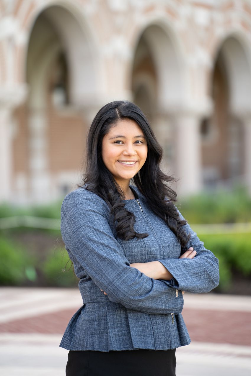A photograph of Esther Jimenez standing outside in front of a building with stone arches that has been blurred. Jimenez has long dark hair and is wearing a blue-grey blouse. Her body is at an angle from the camera and she faces the camera smiling. A photograph of Esther Jimenez standing outside in front of a building with stone arches that has been blurred. Jimenez has long dark hair and is wearing a blue-grey blouse. Her body is at an angle from the camera and she faces the camera smiling.