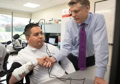 Keith Thomas wears a brain-computer interface with black wires and a medical device on his wrist. He is holding hands with researcher Chad Bouton in a research study room.