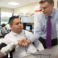 Keith Thomas wears a brain-computer interface with black wires and a medical device on his wrist. He is holding hands with researcher Chad Bouton in a research study room.