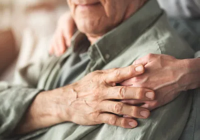 A carer supports the hand of an older man who is seated.