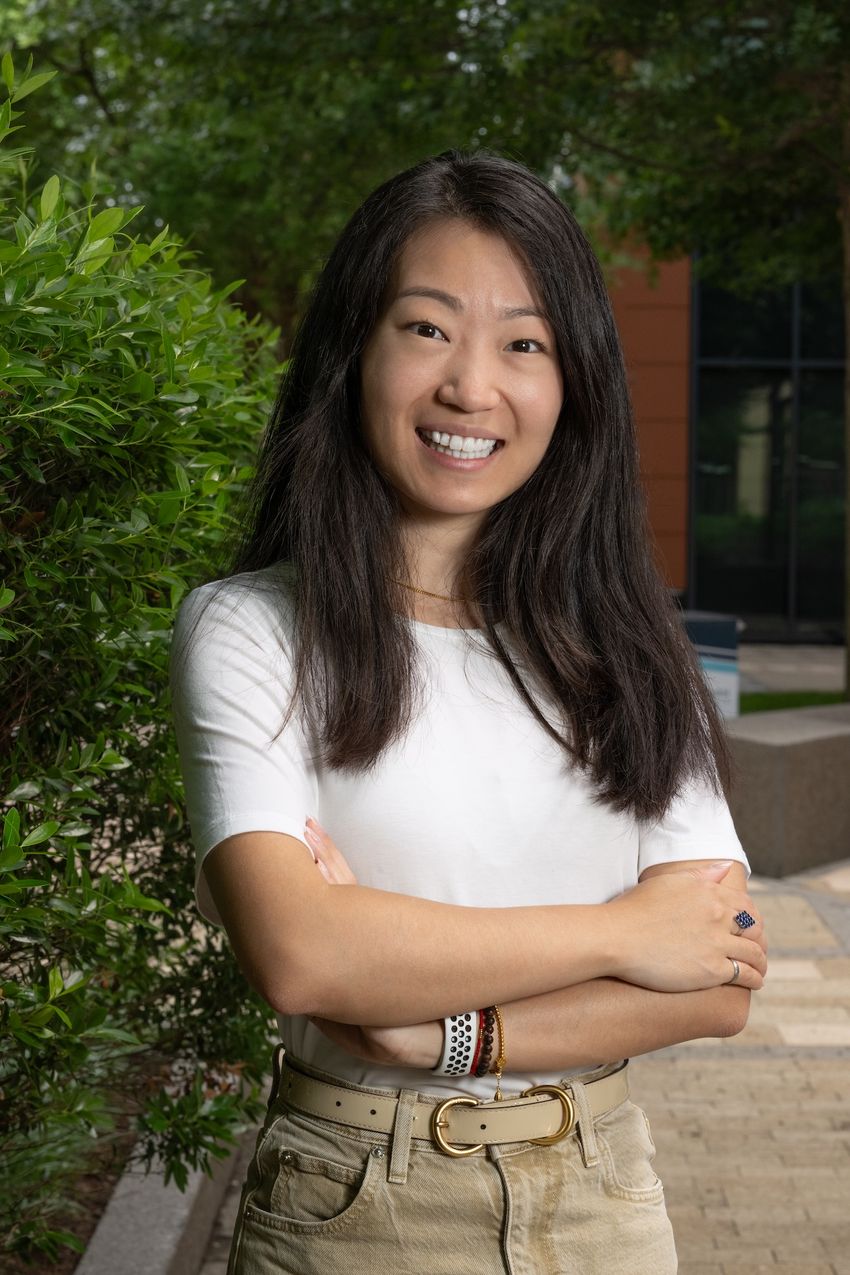 Professional photograph of Jenny Yang standing in front of greenery on a cement patio. Yang has medium-length dark hair and is wearing a white shirt with tan pants. She is crossing her arms and smiling at the camera.