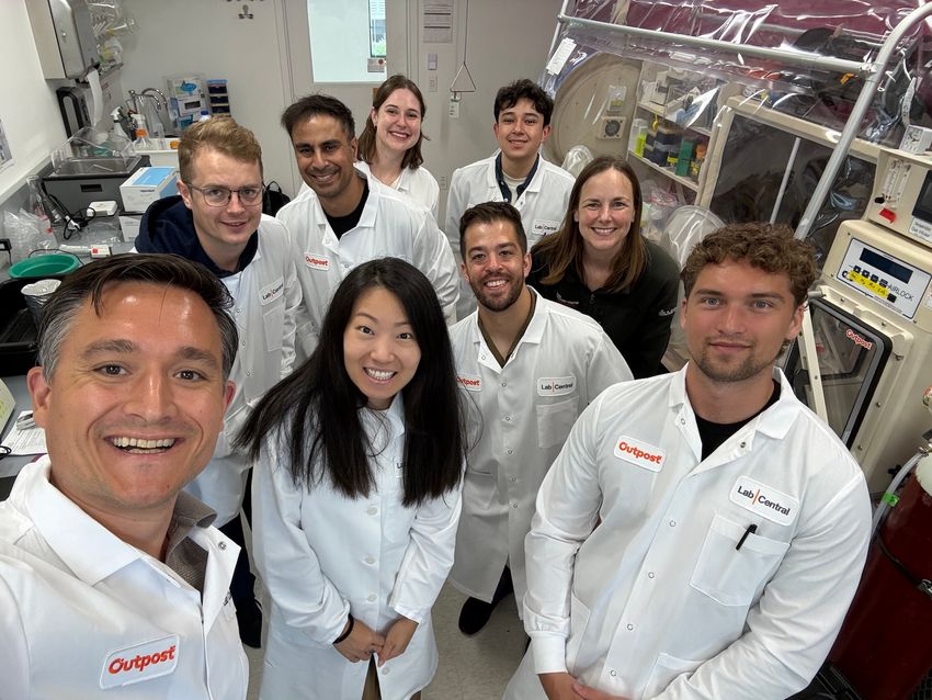 A group of scientists are standing in a laboratory with an anaerobic chamber on the right and a standard lab bench with equipment on the left. Most of the scientists are wearing white lab coats that have a patch that reads “Outpost” in orange letters.