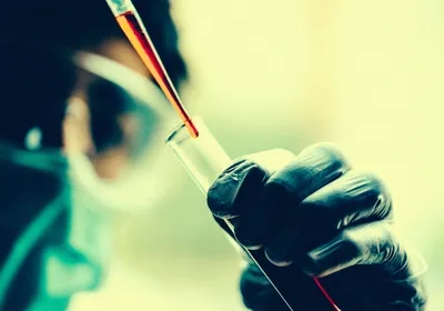 A scientist wearing goggles and a mask pipets red-colored liquid into a tube in a clinical trial setting.