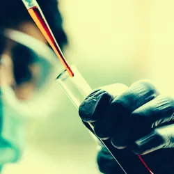 A scientist wearing goggles and a mask pipets red-colored liquid into a tube in a clinical trial setting.