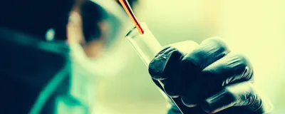 A scientist wearing goggles and a mask pipets red-colored liquid into a tube in a clinical trial setting.