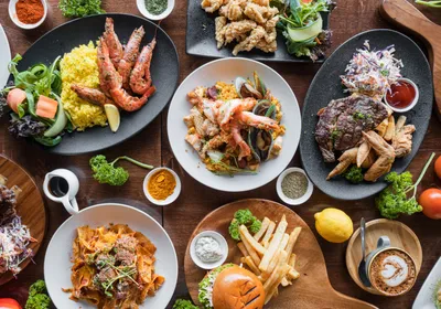 Photograph of a table full of plates of different cuisines, including barbequed meat with coleslaw and fries, seafood with yellow rice and greens, a hamburger and fries, a saucy pasta, and steak with fries.