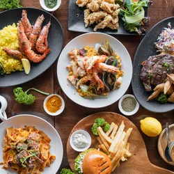 Photograph of a table full of plates of different cuisines, including barbequed meat with coleslaw and fries, seafood with yellow rice and greens, a hamburger and fries, a saucy pasta, and steak with fries. 
