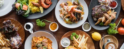 Photograph of a table full of plates of different cuisines, including barbequed meat with coleslaw and fries, seafood with yellow rice and greens, a hamburger and fries, a saucy pasta, and steak with fries. 