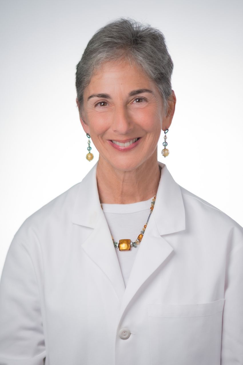 Photograph of Jennifer Grandis, visible from the torso up and standing in front of a white wall. Grandis has short gray hair and is wearing a white lab coat, dangling earrings, and a necklace with orange stones. She is smiling at the camera.