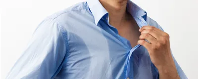 Photograph of a man wearing a light-blue collared shirt, covered in sweat stains at the armpits and chest, standing against a white background. 