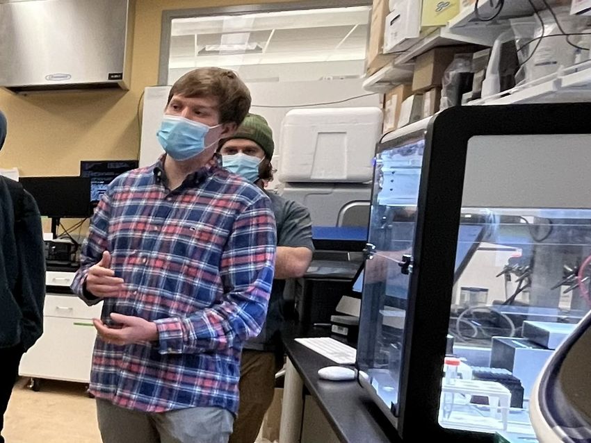 A person wearing a checked shirt and face mask is speaking, standing next to an automated liquid-handling robot. The person standing behind him also wears a face mask.