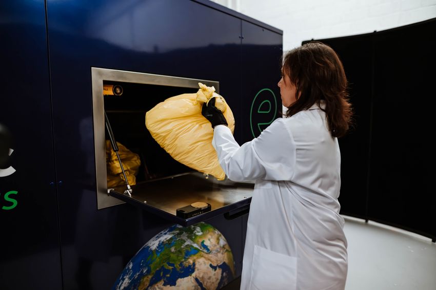 A person wearing a lab coat and gloves puts a yellow-colored bag into a big, blue machine.