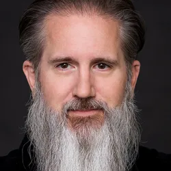Jason T. Gammack has a white beard and looks at the camera against a dark background.