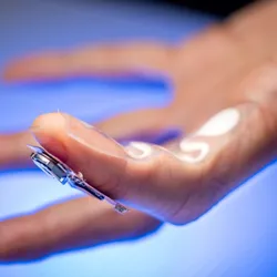 A finger wearing the sweat-sensing device is shown against a bright blue background.