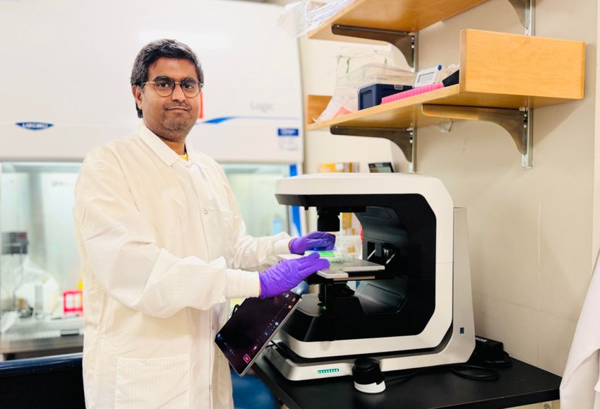 A photograph of Mahesh Chandra Kodali in front of lab equipment. He wears a white lab coat and purple gloves.