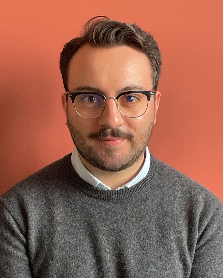 A photograph of Anthony Rabaté wearing a grey sweater and glasses against an orange background.