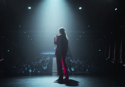 A woman stands in the middle of a stage giving a TED-style talk to an audience.