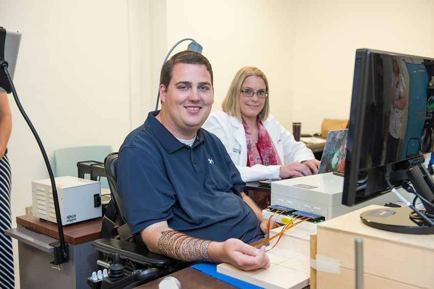 Ian Burkhart sits at a computer with his arm in a sleeve covered in simulators. There is an instrument sitting on his head. A female researcher wearing glasses and a lab coat sits next to him at a computer.