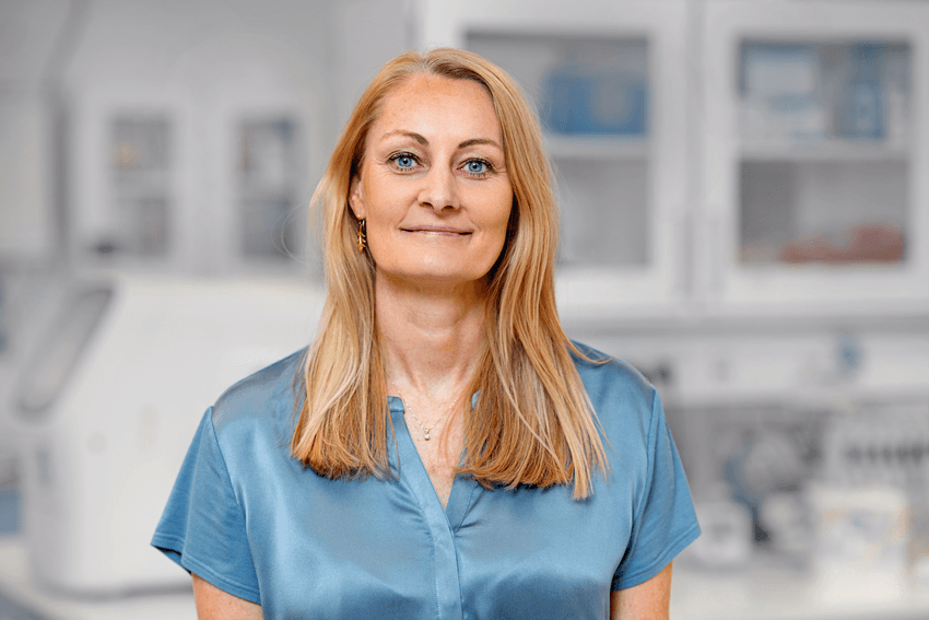 A photograph of Birgitte Rønø standing in a white laboratory, where the background is blurred. Rønø has medium-length blond hair and is wearing a light blue shirt. She is smiling at the camera. A photograph of Birgitte Rønø standing in a white laboratory, where the background is blurred. Rønø has medium-length blond hair and is wearing a light blue shirt. She is smiling at the camera.