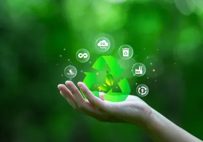Photograph of a person’s hand raised against a background with green, leafy trees. There are icons representing concepts of recycling, reducing carbon dioxide, and reusing materials floating above the hand. 