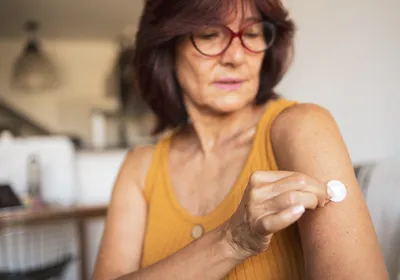 A woman in her 60s wears a yellow tank top and brown-rimmed glasses while placing a hormone-replacement patch on her upper arm.