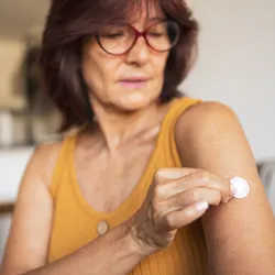 A woman in her 60s wears a yellow tank top and brown-rimmed glasses while placing a hormone-replacement patch on her upper arm.
