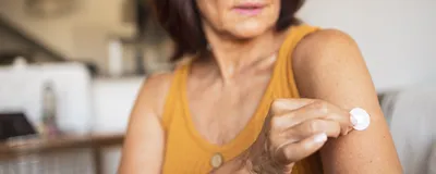  A woman in her 60s wears a yellow tank top and brown-rimmed glasses while placing a hormone-replacement patch on her upper arm.