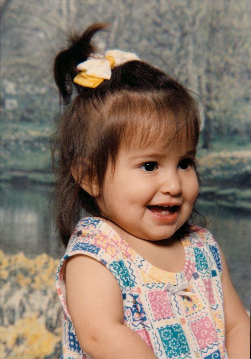 A photograph of Savannah as a toddler. She wears a multicolored top and has a yellow bow in her hair. A photograph of Savannah as a toddler. She wears a multicolored top and has a yellow bow in her hair.