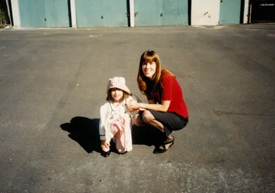 A photograph of a young Savannah and Tracy Dixon-Salazar sitting on the ground. Savannah wears a pink dress and hat and white cardigan, and Tracy wears a red top and black skirt.