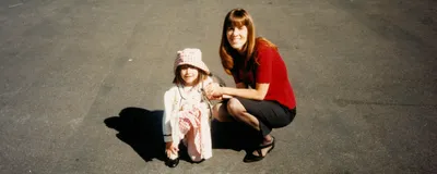 A photograph of a young Savannah and Tracy Dixon-Salazar sitting on the ground. Savannah wears a pink dress and hat and white cardigan, and Tracy wears a red top and black skirt.