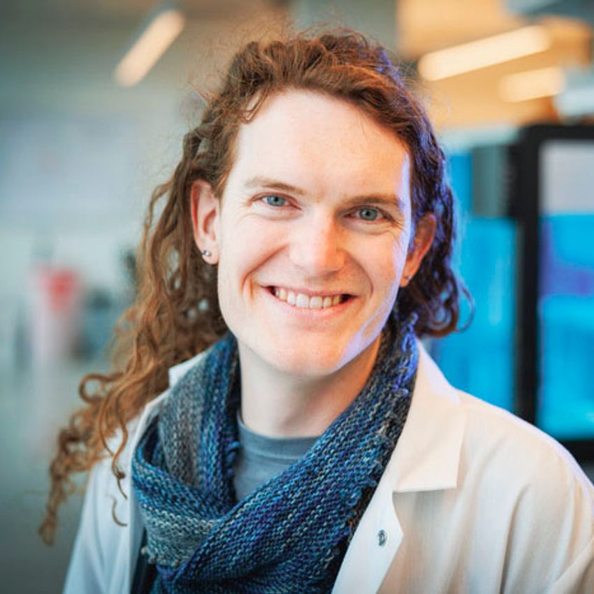 A photograph of Nasa Sinnott-Armstrong standing against a blurred background. Sinnott-Armstrong has long, curly hair and is wearing a white lab coat over a blue and green knitted garment and t-shirt. A photograph of Nasa Sinnott-Armstrong standing against a blurred background. Sinnott-Armstrong has long, curly hair and is wearing a white lab coat over a blue and green knitted garment and t-shirt.