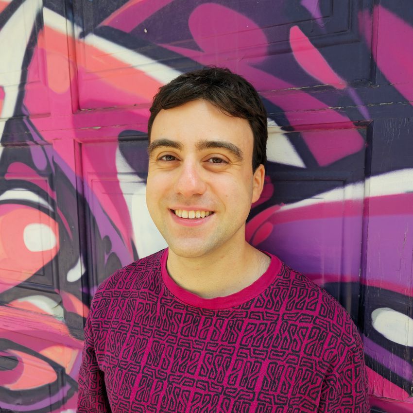 A photograph of Michael Wainberg standing against a purple and pink mural. Wainberg has short, dark hair and is wearing a pink sweater with a dark repeating pattern. A photograph of Michael Wainberg standing against a purple and pink mural. Wainberg has short, dark hair and is wearing a pink sweater with a dark repeating pattern.