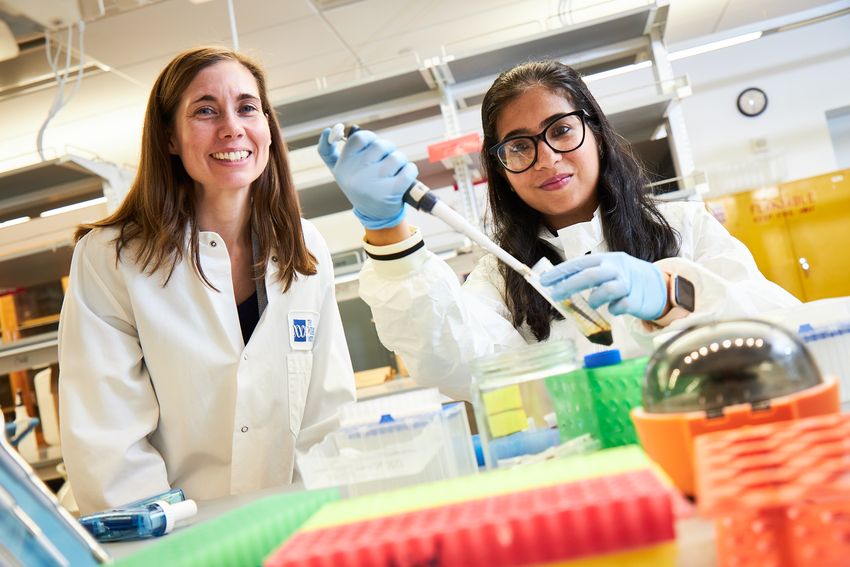 Two women wear white laboratory coats. One is holding a pipette and conical tube.
