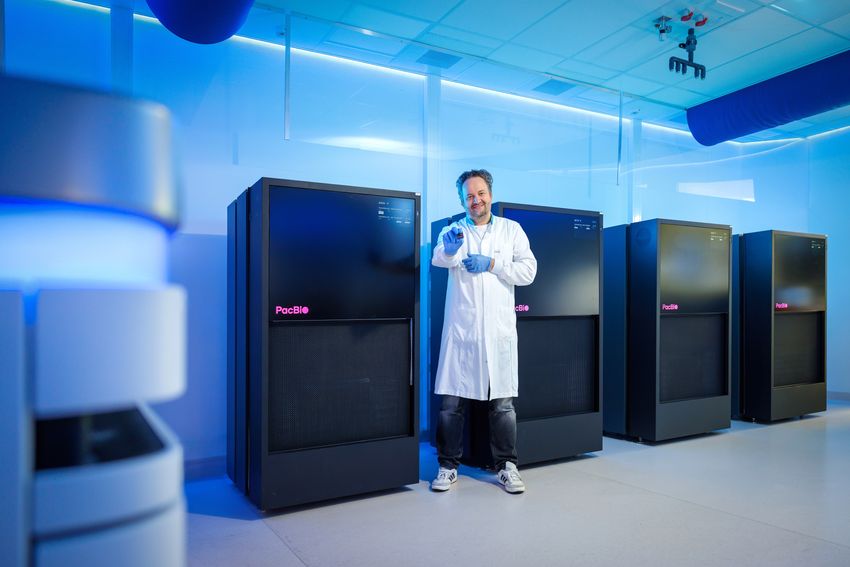 A photograph of Alexander Hoischen wearing a lab coat standing next to a few refrigerator-sized machines. A photograph of Alexander Hoischen wearing a lab coat standing next to a few refrigerator-sized machines.