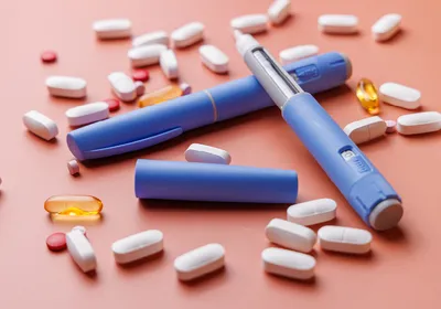 Blue semaglutide GLP-1 drug injectors are shown with other medications for obesity in pill form against a salmon-colored background.