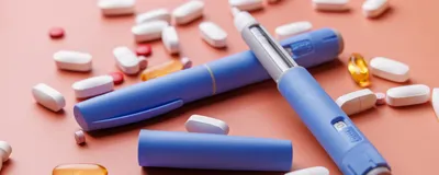 Blue semaglutide GLP-1 drug injectors are shown with other medications for obesity in pill form against a salmon-colored background.