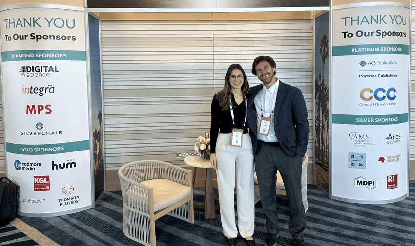 A photograph of Natalie Khalil (left) wearing a dark, long-sleeved top and light pants and her cofounder TJ Reed (right), wearing a light shirt under a dark suit jacket and dark pants. Both are wearing lanyards and standing outside a conference hall.