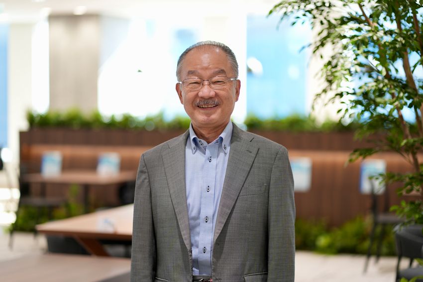 Photograph of Kunihiro Hattori standing in front of a blurred background of tables and indoor plants. He is wearing a grey suit jacket over a light blue collared shirt and has wire-framed glasses. Photograph of Kunihiro Hattori standing in front of a blurred background of tables and indoor plants. He is wearing a grey suit jacket over a light blue collared shirt and has wire-framed glasses.
