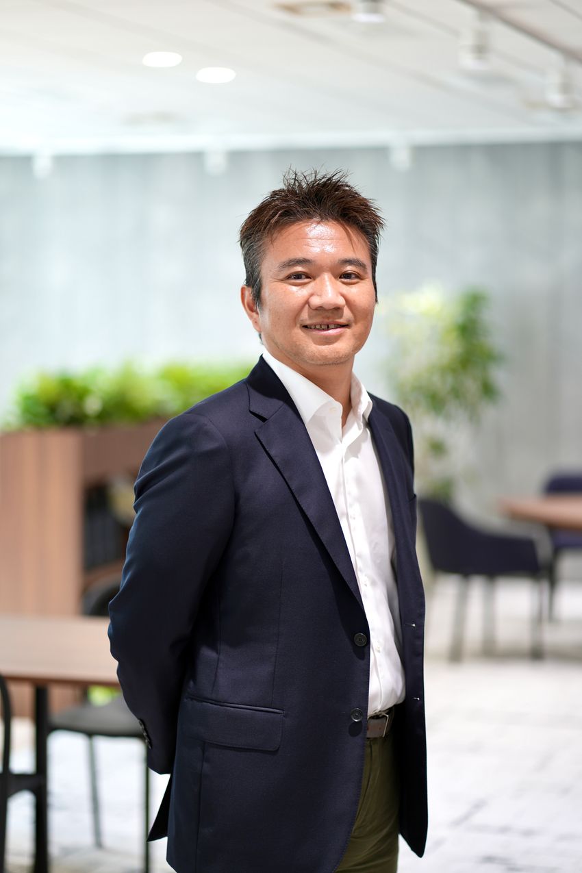 Photograph of Tomoyuki Igawa standing in front of a blurred background of tables and chairs and indoor plants. Igawa is wearing a dark suit jacket over a white collared shirt and has short black hair. Photograph of Tomoyuki Igawa standing in front of a blurred background of tables and chairs and indoor plants. Igawa is wearing a dark suit jacket over a white collared shirt and has short black hair.