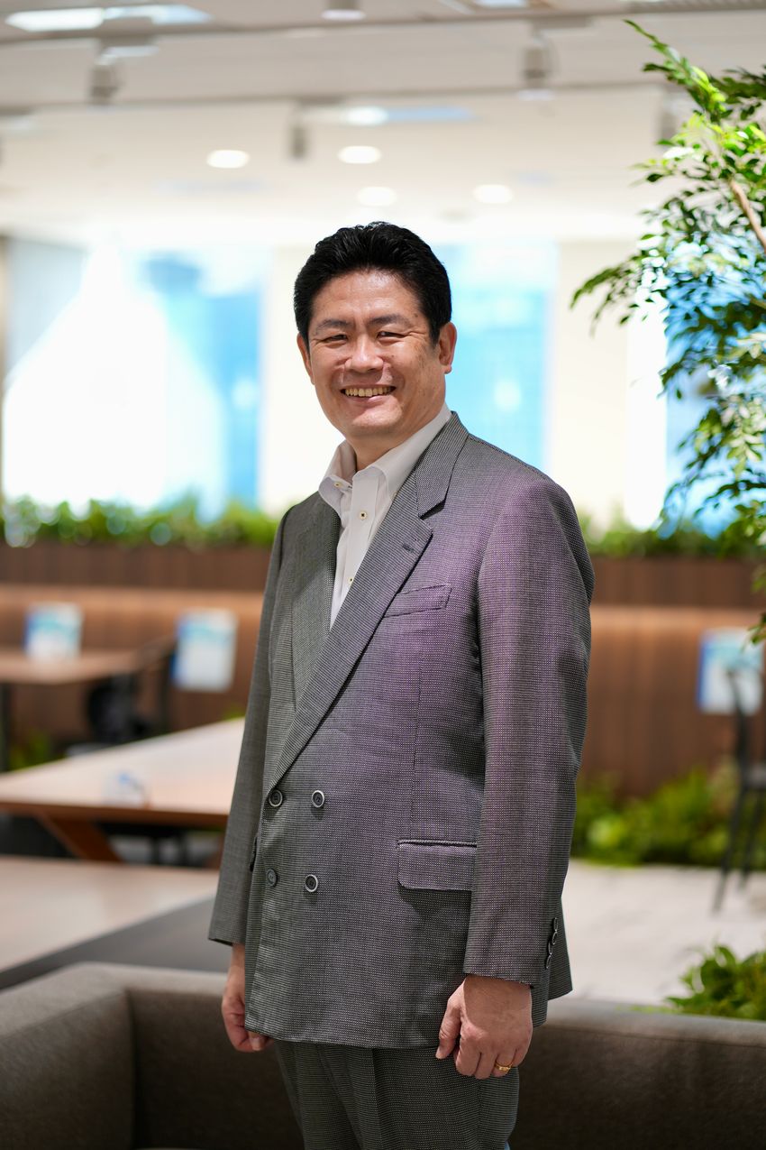 Photograph of Takehisa Kitazawa standing in front of a blurred background of a seating area with indoor plants. He is wearing a grey suit jacket over a light colored, collared shirt and has short black hair. Photograph of Takehisa Kitazawa standing in front of a blurred background of a seating area with indoor plants. He is wearing a grey suit jacket over a light colored, collared shirt and has short black hair.