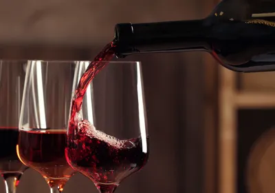 Photograph of wine glasses full of different colored wines. The last glass in the line is being filled with a red wine from a dark bottle. 