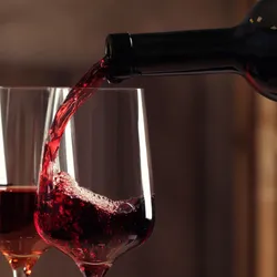 Photograph of wine glasses full of different colored wines. The last glass in the line is being filled with a red wine from a dark bottle. 