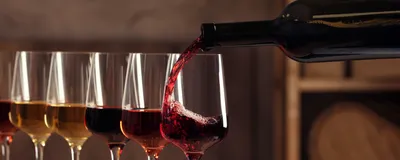 Photograph of wine glasses full of different colored wines. The last glass in the line is being filled with a red wine from a dark bottle. 