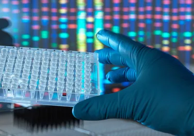 A scientist wearing a blue glove holds a 96-well plate in their hand. Metagenomic data is shown on the computer screen in the background.