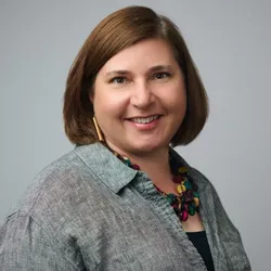 Kate Travis wears a grey shirt and colorful necklace in a headshot.