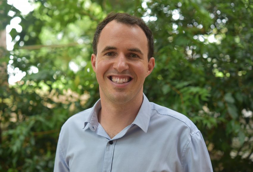 Seth Cheetham smiles, wearing a light blue button-down shirt, against a green leafy backdrop.